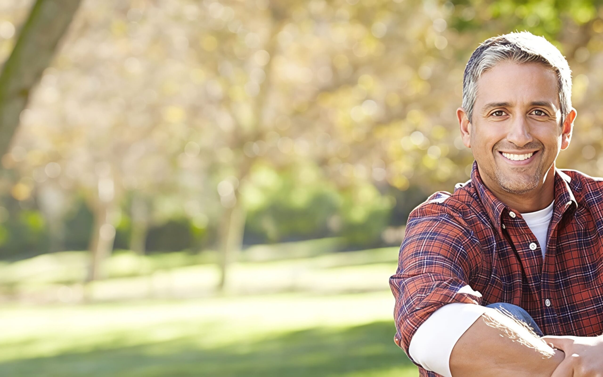A man with a plaid shirt smiling at the camera. Grass and trees blurred in the background.