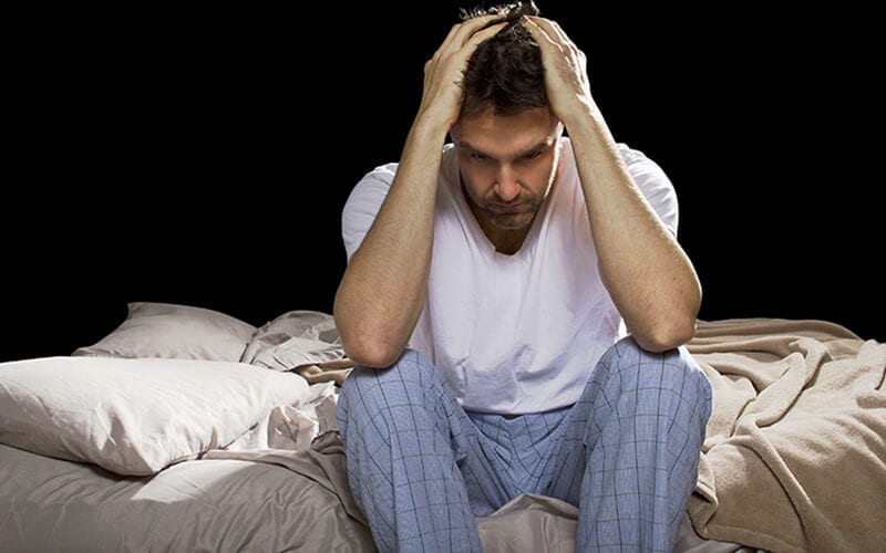 A man in Pajamas sitting up in bed with hands holding his head.