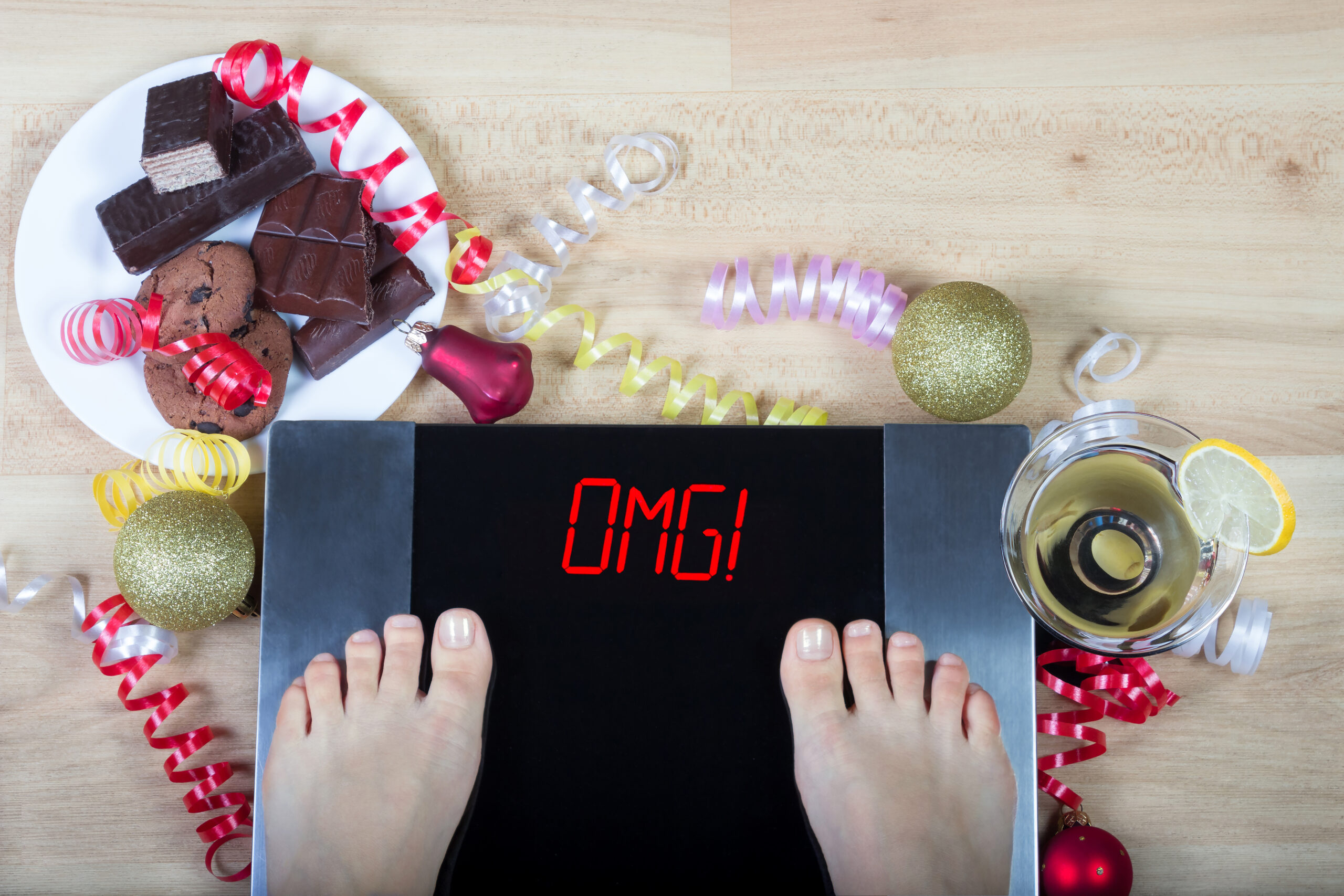 A person standing on a scale, which reads OMG. Around the scale is party supplies, a martini, and chocolate.