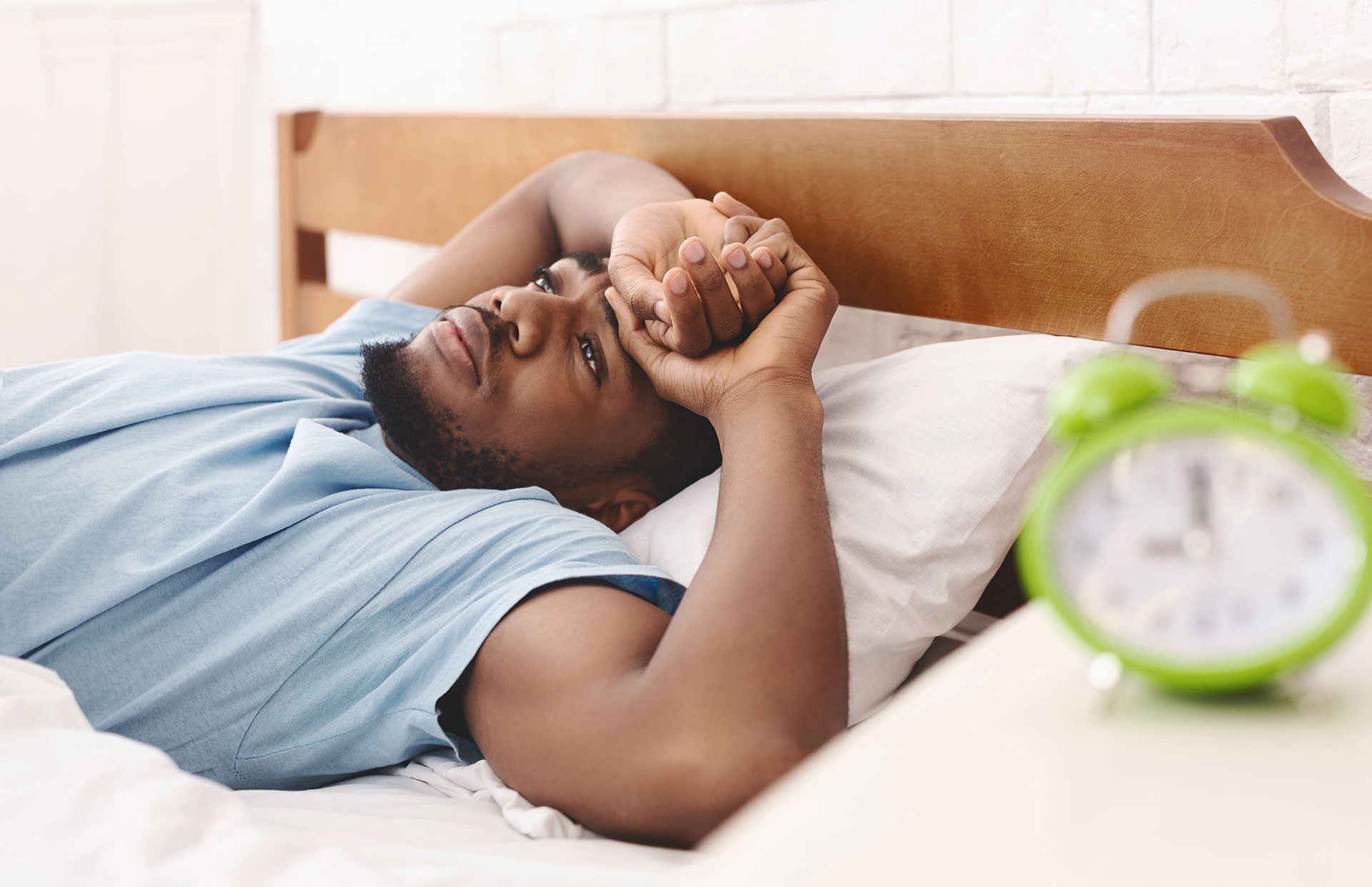 A disconcerted man laying in bed looking at the ceiling. a green alarm clock on a nightstand next to the bed.