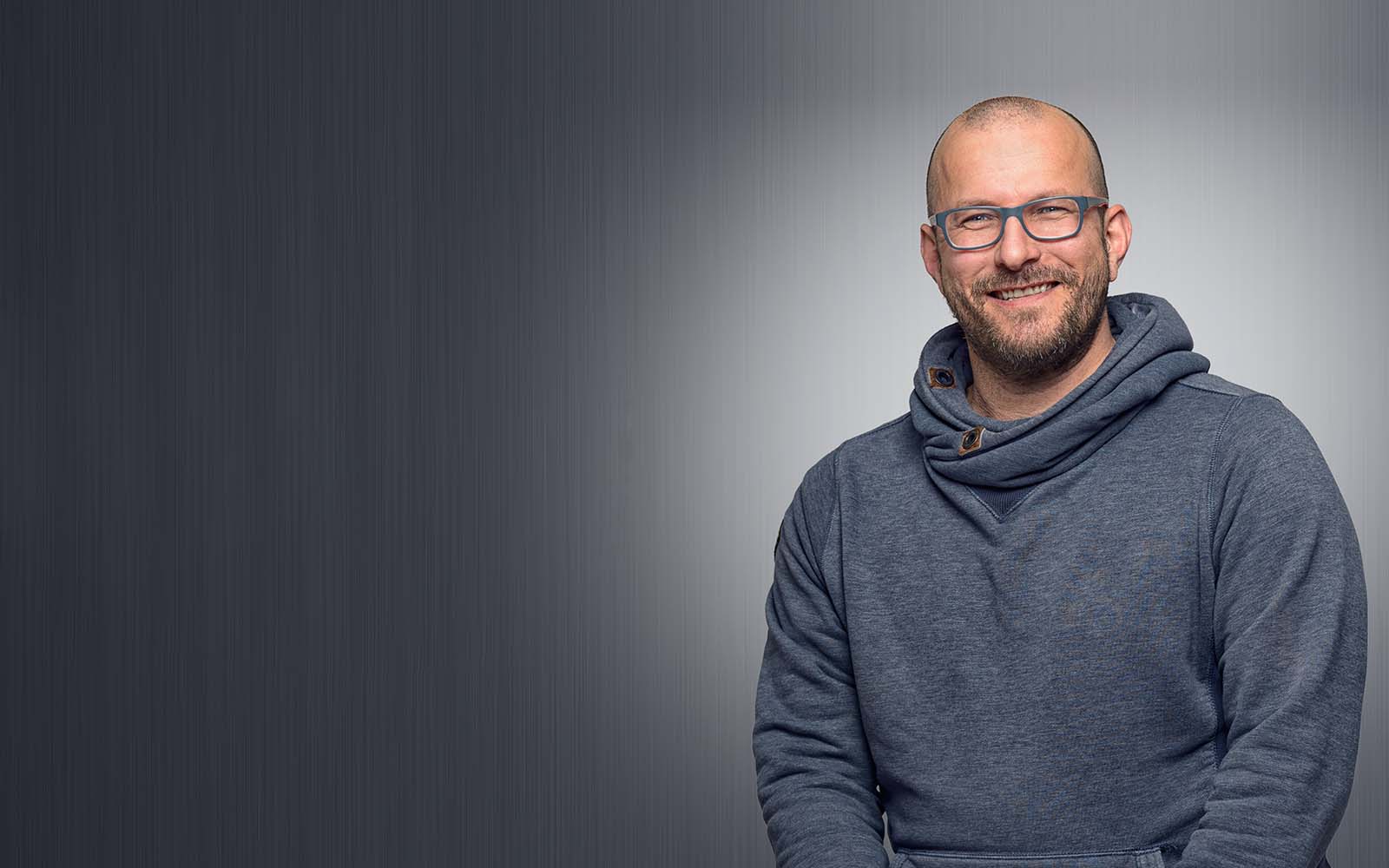A male in a sweater with glasses, smiling on a studio background.